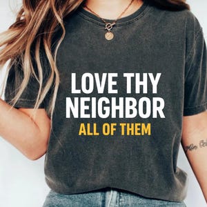 Peut inclure: T-shirt gris foncé avec l'inscription blanche et jaune "LOVE THY NEIGHBOR ALL OF THEM". Le t-shirt est de style col rond et est porté avec un jean bleu. Le modèle porte un collier doré.