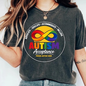 Puede incluir: Camiseta gris oscuro con un diseño gráfico circular que promueve la aceptación del autismo. El diseño presenta un símbolo de infinito arcoíris, la palabra "AUTISM" en colores del arcoíris y el texto "Acceptance" y "Proud Autism Mom".
