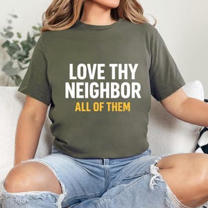 Può includere: T-shirt verde oliva con la scritta "LOVE THY NEIGHBOR ALL OF THEM" in bianco e giallo. La maglietta è indossata con jeans blu. Il tema generale è un messaggio di inclusione e gentilezza.