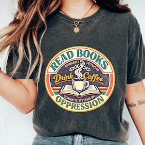 Peut inclure: T-shirt gris foncé avec un motif circulaire. Le motif indique : « Lire des livres, démanteler les systèmes d'oppression, boire du café ». Le motif comprend une tasse à café, un livre ouvert et des couleurs rétro vertes, rouges, jaunes et oranges.