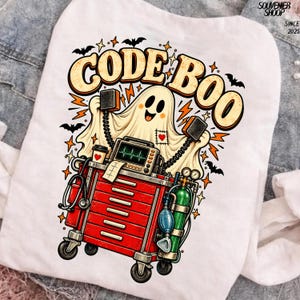 Puede incluir: Una camiseta blanca con un gráfico de un fantasma detrás de un carro médico rojo. El fantasma sostiene dos paletas de desfibrilador. El carro incluye un monitor cardíaco, un estetoscopio y un tanque de oxígeno. Texto CODE BOO arriba.