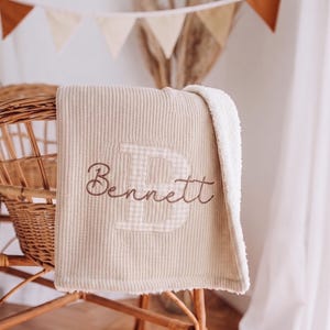 May include: A beige waffle-knit blanket with a white sherpa fleece backing. The blanket features a large, light-coloured gingham letter B embroidered in the centre, with the name Bennett stitched in brown cursive script across the letter.
