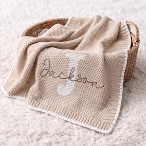 May include: A beige knitted blanket with a textured waffle pattern and a white border. The blanket features a large, light-coloured striped letter J with the name Jackson embroidered in brown cursive script across the centre of the letter.