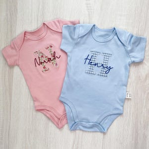 May include: Two baby bodysuits, one pink and one light blue. The pink bodysuit has a floral "N" with the name "Norah" in script. The blue bodysuit features a gingham "H" with the name "Henry" in script. Both are short-sleeved.