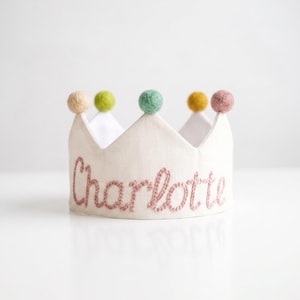 May include: A white fabric crown featuring the name Charlotte embroidered in pink cursive thread. The top edge of the crown is decorated with five small, round, felted pom-poms in shades of beige, light green, mint green, mustard yellow, and dusty pink.