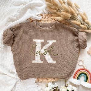 May include: A brown knit sweater with the name "Kash" embroidered in green cursive script over a large letter "K" with a gingham pattern. The sweater is displayed on a woven mat with decorative elements.