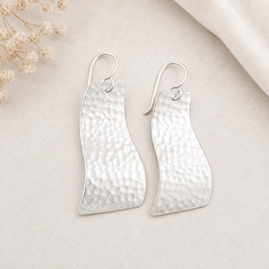 May include: A pair of silver-colored, hammered-texture earrings. The earrings have a curved, rectangular shape with a hook closure. The background is a neutral color with a white cloth and small white flowers.