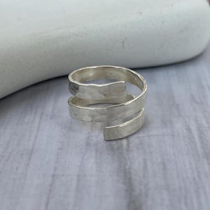 Op de afbeelding: Een zilverkleurige wikkelring met een gehamerde textuur. De ring heeft een spiraalvormig ontwerp, met twee volledige wikkelingen en een open uiteinde. De ring wordt getoond op een witte steen tegen een lichtgrijze houten ondergrond.