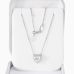 May include: A silver-toned box chain necklace featuring a heart-shaped pendant with a central clear stone surrounded by a halo of smaller stones. The necklace includes an adjustable sliding clasp mechanism with a small heart-shaped charm at the end.