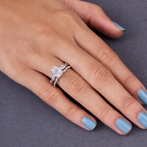 Peut inclure: Une bague argentée avec une pierre centrale ovale transparente montée sur griffes, accompagnée de deux anneaux entrelacés incrustés de petites pierres transparentes rondes qui s'étendent le long de l'anneau de la bague.