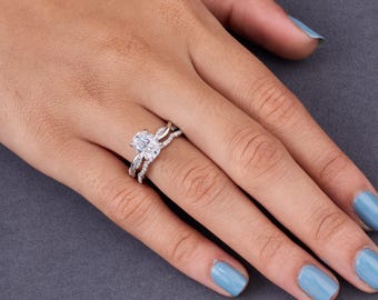 Pode incluir: Um anel de tom prateado com uma pedra central transparente de corte oval em cravação de garras, ladeado por duas bandas entrelaçadas incrustadas com pequenas pedras redondas e transparentes ao longo do aro do anel.