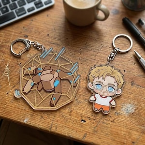 May include: Two acrylic keychains with silver keyrings. One features a brown and blue robot design within a geometric shape. The other depicts a cartoon character with glasses, a white shirt, and orange shorts.
