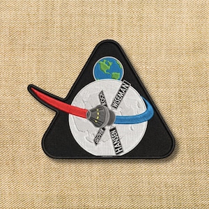 May include: Embroidered patch featuring a black triangle with a stylized Earth and a white circle depicting the moon. A red and gray spacecraft extends from the center. Names of astronauts are printed on the circle.