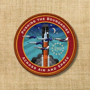 May include: Embroidered patch with a rocket launch scene, fighter jets, and the text "PUSHING THE BOUNDARIES ACROSS AIR AND SPACE." The patch is circular with a red border and features a blue and orange color scheme.