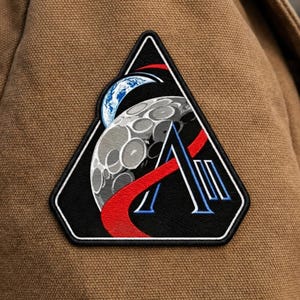May include: A black and white patch with a space-themed design. The patch features a stylized moon, Earth, and the letter "A" with red and blue accents. The patch is sewn onto a brown fabric.