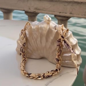 Peut inclure: Un sac à main en forme de coquillage nacré, avec une chaîne dorée et un foulard décoratif. Le sac est orné de deux perles. En arrière-plan, une scène de bord de mer avec des bâtiments et des gondoles.