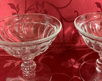 May include: Two clear glass dessert bowls with a stemmed base. The bowls have a scalloped design around the rim and a spiral design on the stem. The bowls are on a red background.
