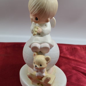 May include: A porcelain figurine of a little angel with blonde hair and a halo, sitting on a globe with a teddy bear sitting on a box in front of her. The figurine is white with pink accents.