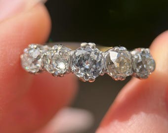 Könnte beinhalten: Nahaufnahme eines Rings mit fünf klaren, facettierten Edelsteinen, die in einem silbernen Band gefasst sind. Die Edelsteine sind rund und gleichmäßig angeordnet, mit einer leichten Farbvariation. Der Ring wird zwischen zwei Fingern gehalten.