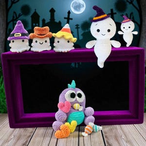 May include: A collection of handmade crochet Halloween decorations. The image features white ghost figures wearing hats, a purple bird-like creature, and colorful candies. The decorations are arranged on a purple frame with a spooky background.