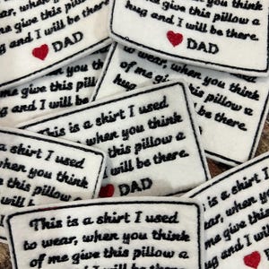 Puede incluir: Parches rectangulares blancos con texto bordado en negro y un corazón rojo. El texto dice: "Esta es una camisa que solía usar, cuando pienses en mí dale un abrazo a esta almohada y estaré allí. PAPÁ."