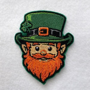 Puede incluir: Parche bordado con un duende con sombrero verde, cinturón negro y hebilla naranja. El duende tiene barba roja y expresión sonriente. Un trébol verde está en el sombrero. El parche tiene un borde verde.