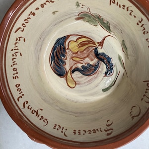 May include: A ceramic bowl with a cream-colored interior and a reddish-brown rim. The bowl features a painted design of a fruit with blue, yellow, and brown details, along with green leaves. The rim has handwritten text in a dark brown color.