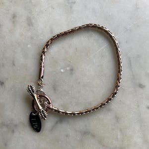May include: A rose gold bracelet with a woven chain design. The bracelet features a toggle clasp and a small tag that reads "7.75". The bracelet is displayed on a marble surface.