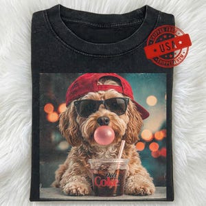 May include: A black crew neck t-shirt featuring a graphic of a light brown dog wearing a red baseball cap and black sunglasses. The dog is blowing a pink bubble and holding a clear plastic cup with a straw and the red text Coke printed on it.