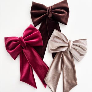 May include: Three velvet bows in shades of brown, burgundy, and taupe. Each bow features a large, looped design with long, flowing tails. The bows are arranged against a white background, highlighting their rich colors and textures. These accessories are suitable for hair or gift wrapping.