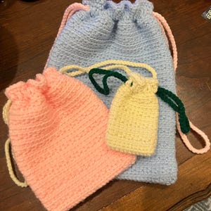 Puede incluir: Tres bolsas de crochet hechas a mano con cordón en colores pastel. Una bolsa grande azul claro con cordón rosa, una bolsa melocotón mediana con cordón amarillo y una bolsa amarilla pequeña con cordón verde. Las bolsas están hechas de hilo suave.