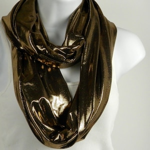 May include: A gold metallic infinity scarf draped around a white mannequin. The scarf has a shiny, reflective surface and is made of a soft, flowing material. The scarf is a solid color and appears to be made of a lightweight fabric.
