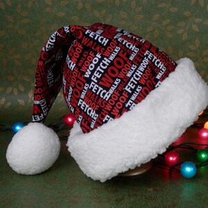 Puede incluir: Un gorro de Papá Noel rojo y negro con ribete de piel blanca. El gorro está cubierto con las palabras "Woof Walks Fetch" repetidas en un patrón.