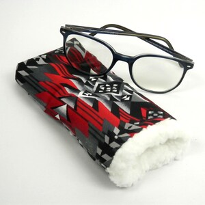 May include: Eyeglasses rest on a patterned case. The case features a geometric design in red, black, gray, and white. The interior of the case is lined with white, fluffy material. The eyeglasses have dark blue frames.