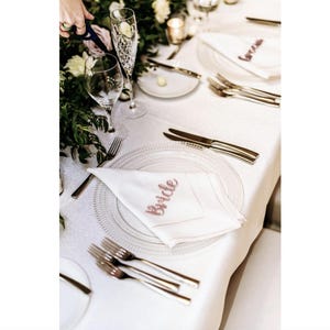 May include: A wedding reception table setting with white linens, clear glass plates, and silverware. White napkins with the word "Bride" in pink script are folded on the plates. Champagne flutes and wine glasses are also present.