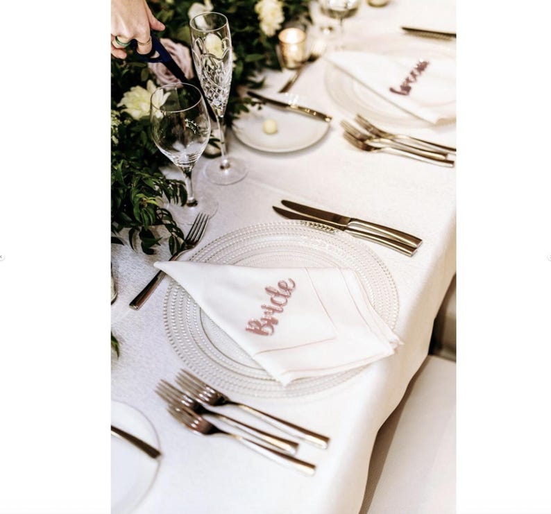 May include: A wedding reception table setting with white linens, clear glass plates, and silverware. White napkins with the word "Bride" in pink script are folded on the plates. Champagne flutes and wine glasses are also present.