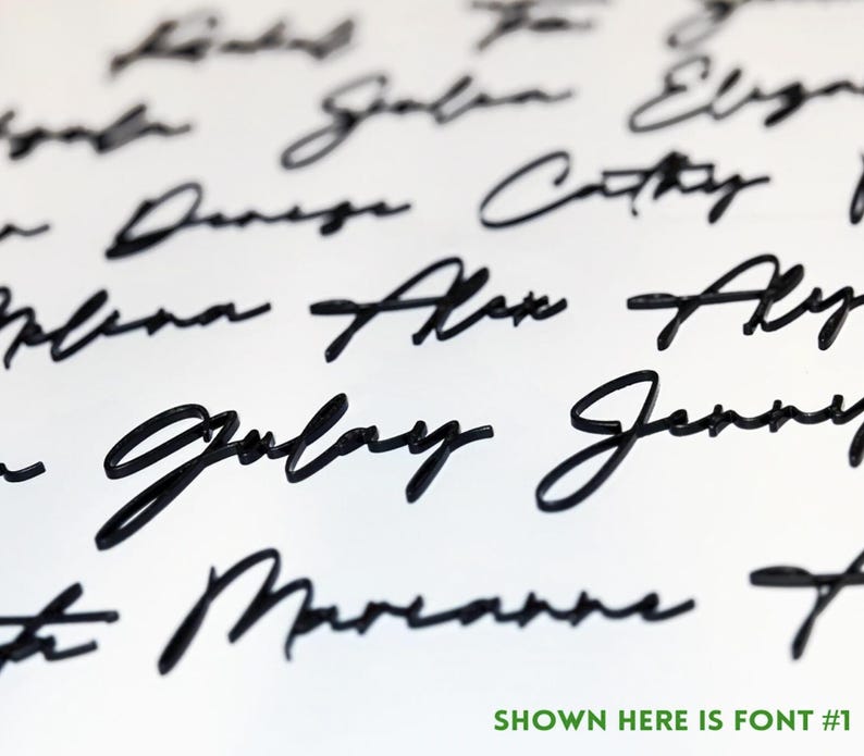 May include: Close-up of a white surface with multiple names written in black cursive script. The text "SHOWN HERE IS FONT #1" is in green at the bottom. The names are in a flowing, elegant font.