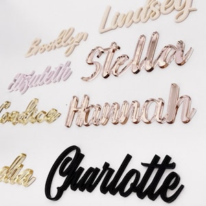 May include: A collection of custom name cutouts in various colors and fonts. Names include "Lindsey", "Brooklyn", "Elizabeth", "Candice", "Hannah", and "Charlotte". The names are in gold, rose gold, and black.