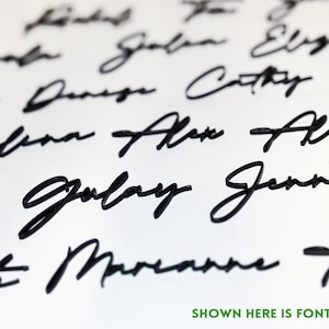 May include: Close-up of a white surface with multiple names written in black cursive script. The text "SHOWN HERE IS FONT #1" is in green at the bottom. The names are in a flowing, elegant font.