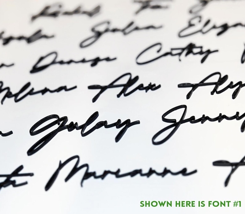 May include: Close-up of a white surface with multiple names written in black cursive script. The text "SHOWN HERE IS FONT #1" is in green at the bottom. The names are in a flowing, elegant font.