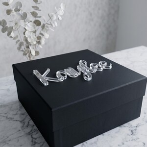 May include: A black gift box with the name "Kaylee" in clear acrylic letters on top. The box is sitting on a marble surface, with a vase of eucalyptus in the background.