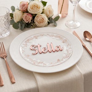 May include: A place setting with a white plate, a clear plate with a floral design, and a rose gold nameplate that says "Stella". A fork, knife, and spoon are on the table. A bouquet of roses and greenery is in the background.