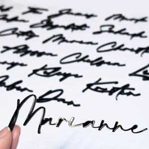 May include: Black cursive name cutouts, with one held by a hand. The name "Marianne" is in focus, with other names blurred in the background. The names appear to be made of a shiny, reflective material, possibly acrylic or metal.