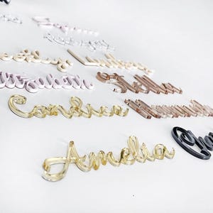 May include: A collection of acrylic name cutouts in various colors, including clear, gold, pink, and black. The names are displayed in a cursive font, perfect for personalized gifts or event decor. The names are scattered on a white surface.