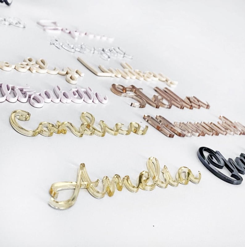 May include: A collection of acrylic name cutouts in various colors, including clear, gold, pink, and black. The names are displayed in a cursive font, perfect for personalized gifts or event decor. The names are scattered on a white surface.