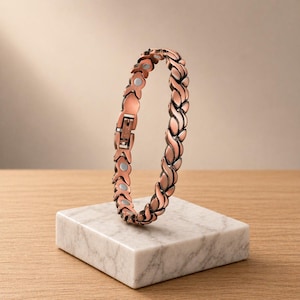 May include: A copper-colored bracelet with a braided design, featuring a clasp closure. The bracelet is displayed on a white marble block, set against a neutral background. The bracelet's links have a leaf-like pattern.