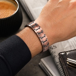 May include: A copper-colored bracelet with a rectangular link design is worn on a wrist. The bracelet has a metallic sheen and a segmented appearance. The wrist is partially visible, and the bracelet is the focal point of the image.