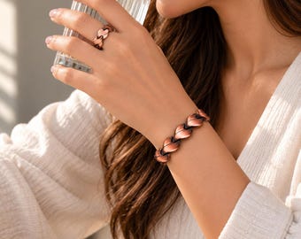 May include: A copper-toned metal bracelet and a matching ring, both featuring a repeating leaf-like link design with dark, oxidized accents between each segment. The jewelry pieces have a polished finish and a structured, articulated construction.
