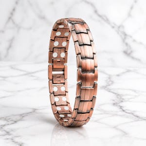 May include: A copper-colored magnetic bracelet with a segmented design. The bracelet features rectangular links and white circular accents. The links are connected to form a flexible band. The bracelet is displayed against a marble background.