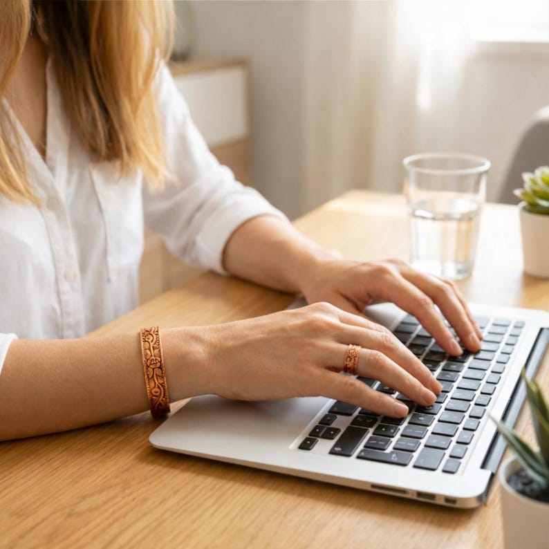 May include: Copper-colored bracelet and ring with floral patterns. The jewelry is worn on a person's hand while they are using a laptop. The bracelet is approximately 0.75 inches wide. The ring has a similar design.